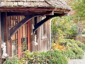 A rustic wooden building with a sloped roof, a bell hanging from the eave, and a statue on the left side.