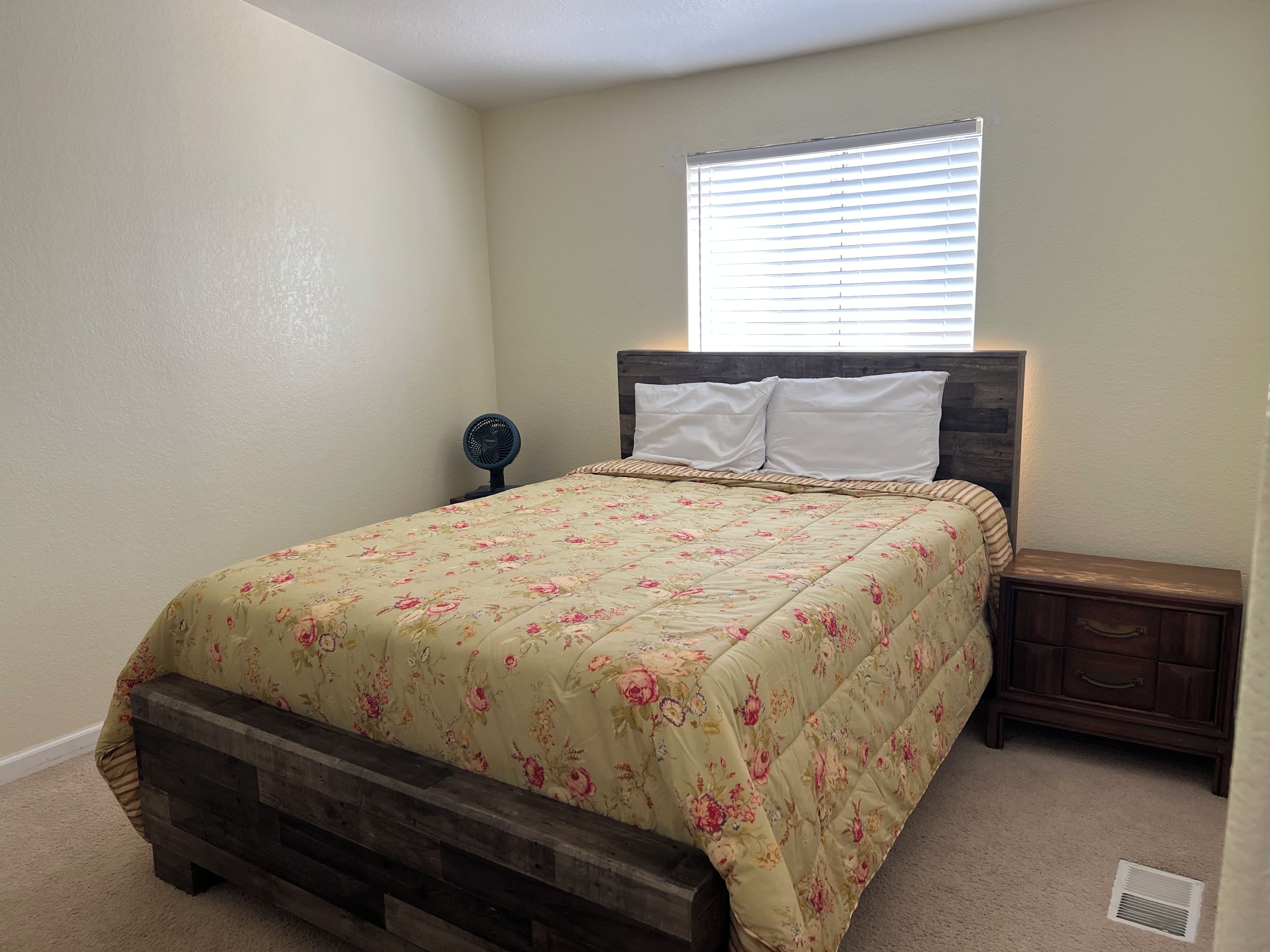 Quarto com cama, mesa de cabeceira, ventoinha na parede e janela com persianas.
