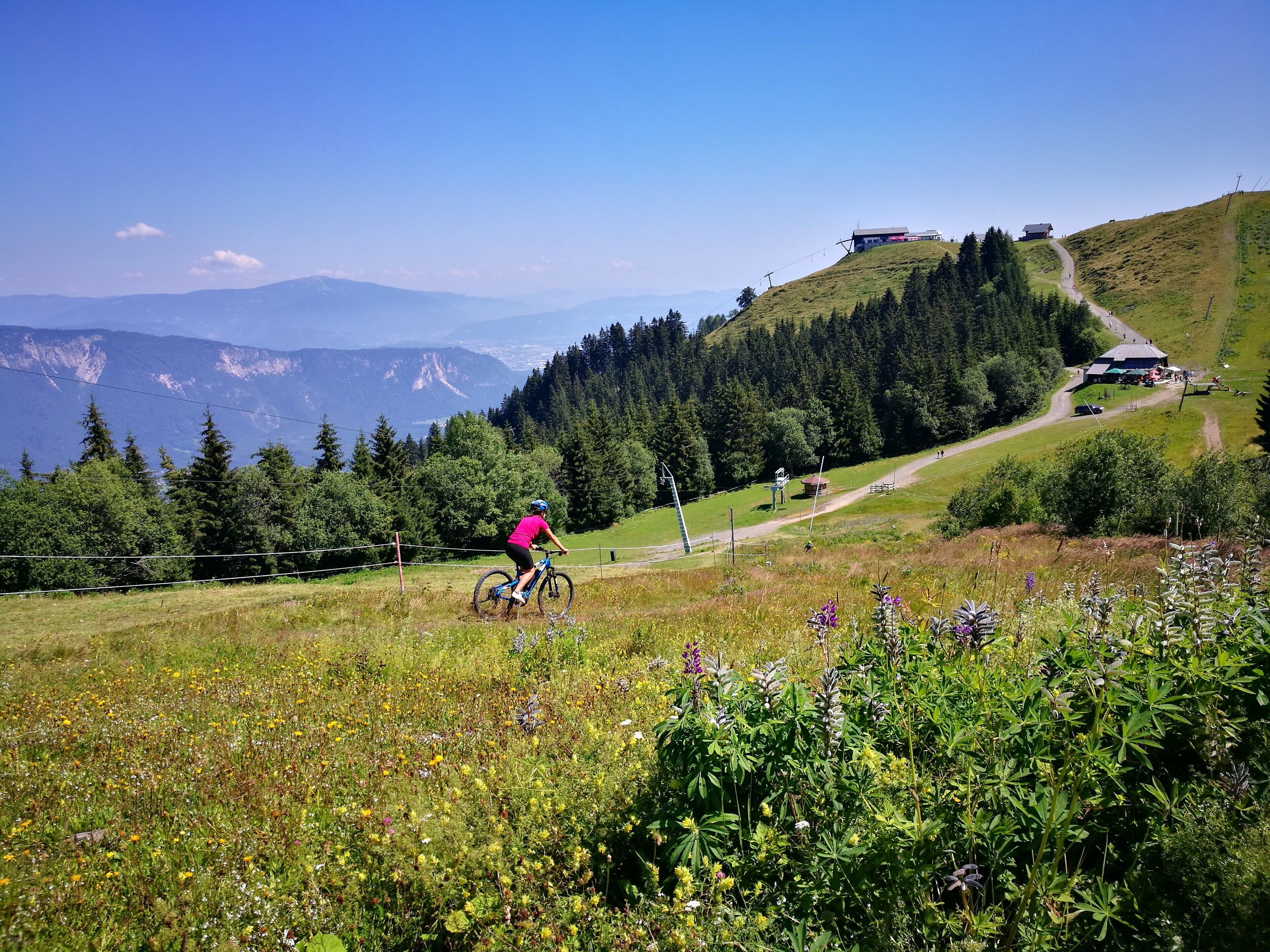 A cyclist on a mountain trail with a view of a mountain range and a building on a hill.