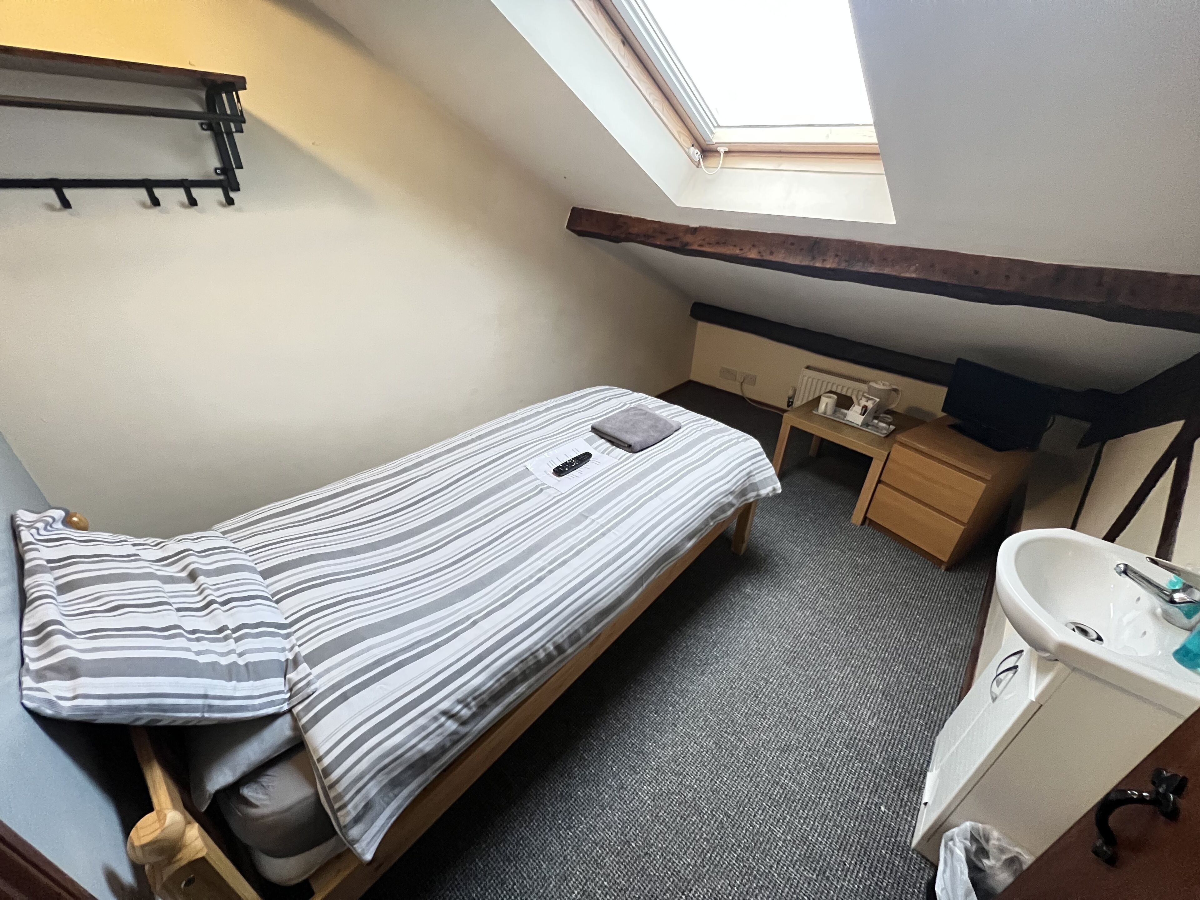 A small, single-bed room with a skylight, a bedside table, a small desk, and a sink.