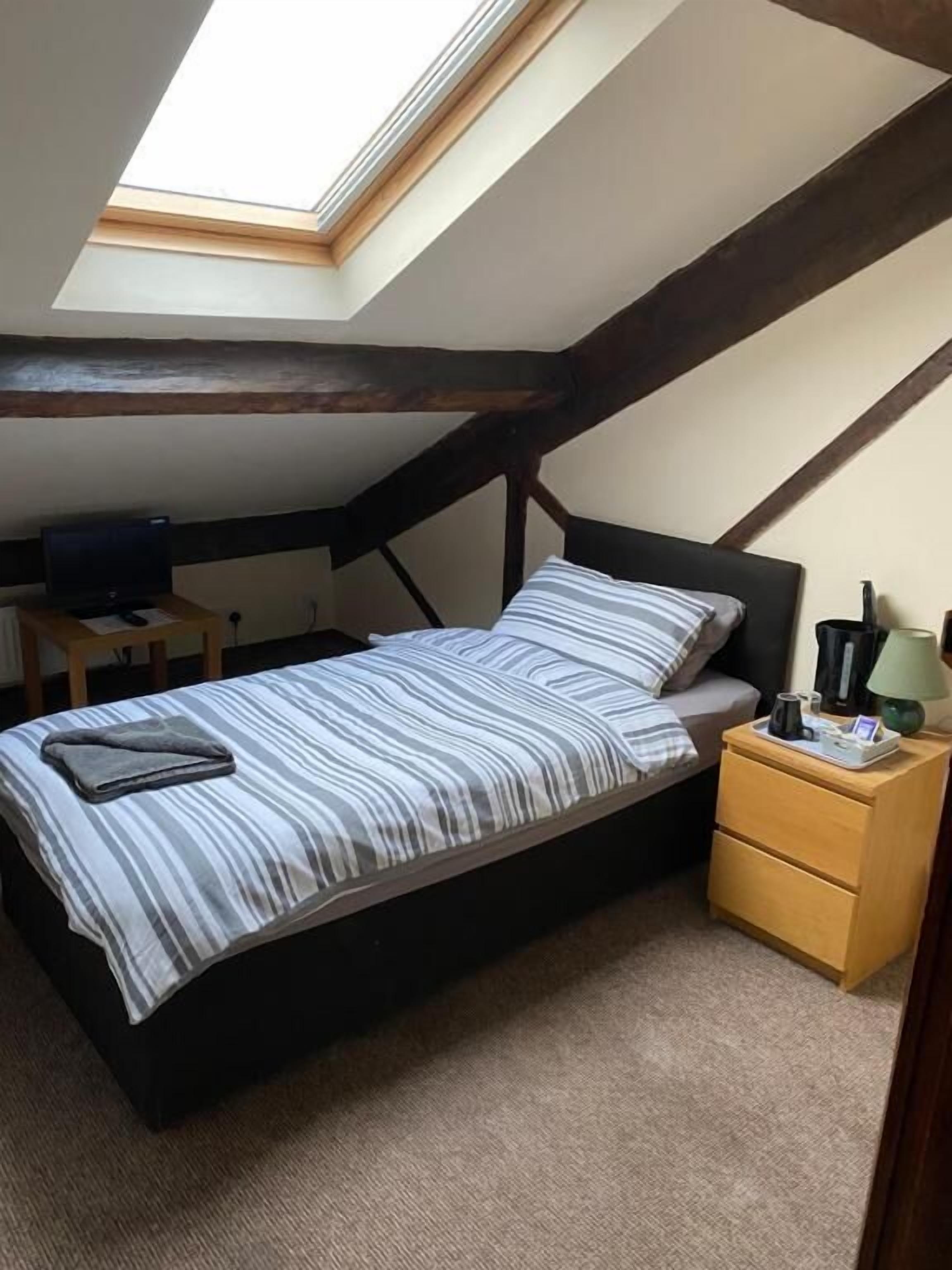 A bedroom with a sloped ceiling, a bed with striped bedding, a bedside table with a lamp, and a small desk with a computer.