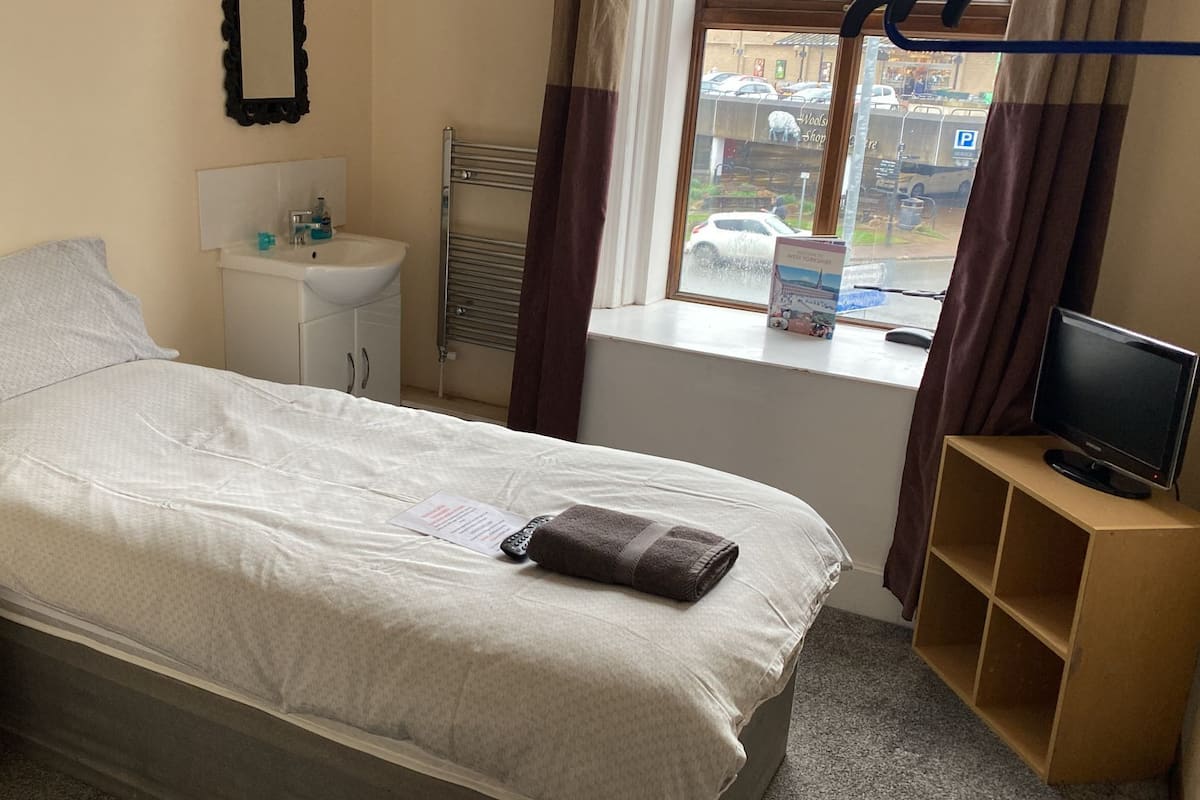A hotel room with a bed, a bedside table, a TV, a window with curtains, and a view of the street outside.