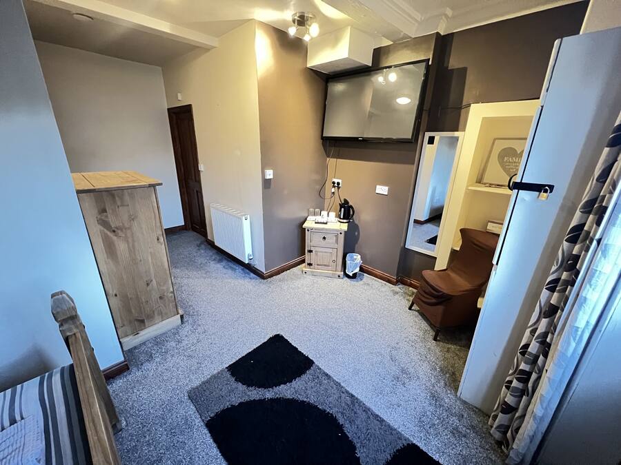 A hotel room with a wooden wardrobe, a small bedside table with a lamp, a radiator, a mirror, and a brown chair.