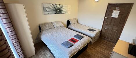 Una habitación con dos camas, piso de madera, una lámpara fijada a la pared y una puerta con un letrero.