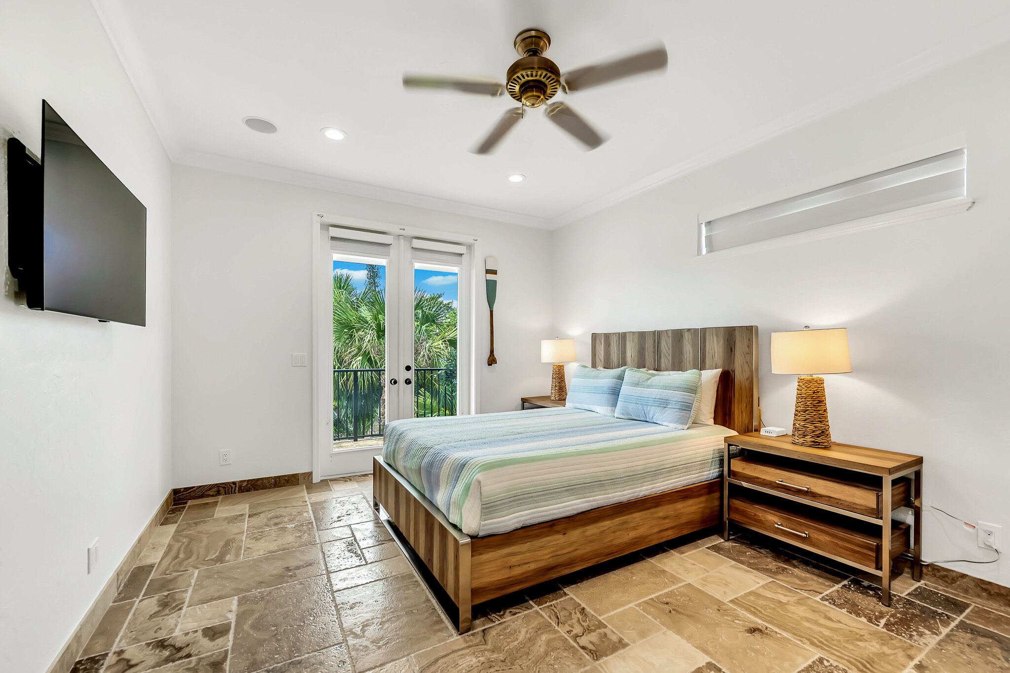 A bedroom with a bed, nightstands, a ceiling fan, a TV, and a view of palm trees through the window.