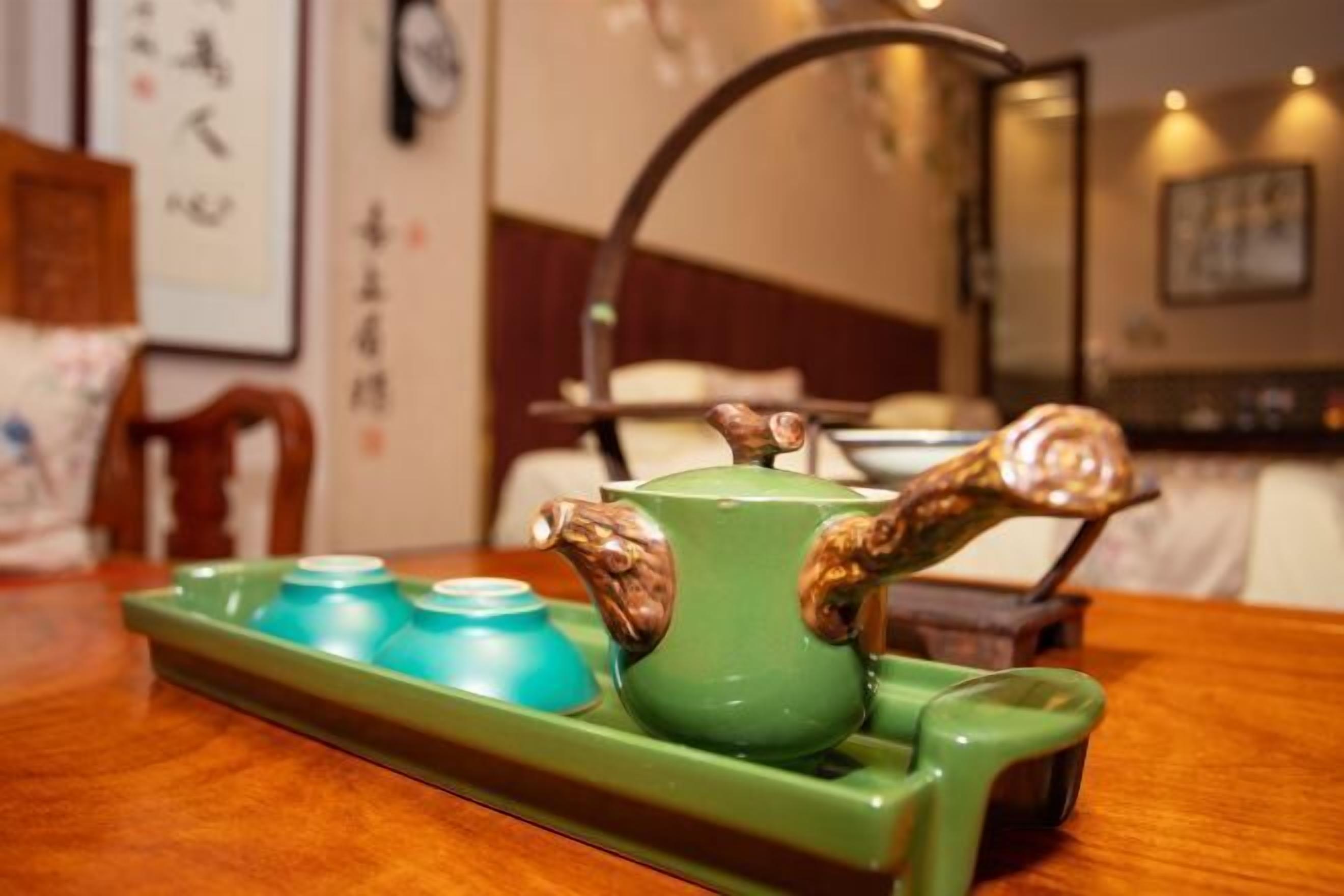 Una tetera verde con motivos de animales sobre una bandeja, acompañada de dos tazas azules, colocadas sobre una mesa de madera en una habitación con una cama y una mesita de noche al fondo.