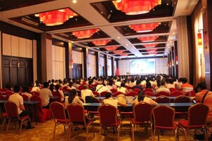 A conference room with rows of tables and chairs, large windows, and a stage at the front.