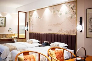 Quarto de hotel com duas camas, cabeceira de madeira, espelho fixado na parede, mesa de cabeceira com abajur e uma pintura decorativa.