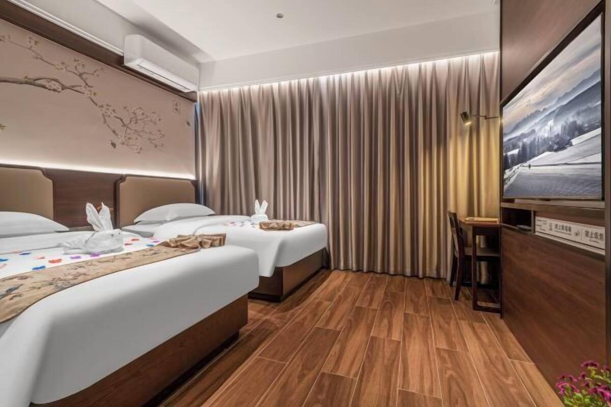 Habitación de hotel con dos camas, suelo de madera, un ventanal grande con cortinas y un televisor de pantalla plana en la pared.