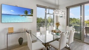 Un comedor moderno con una mesa de vidrio, sillas blancas y un televisor grande mostrando una escena de playa.