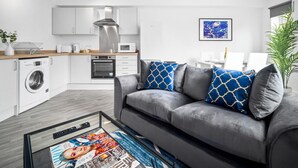 Eine moderne Wohnung mit grauem Sofa, einem Couchtisch mit einer Zeitschrift, einer Waschmaschine, einer Mikrowelle und einem Essbereich mit Tisch und Stühlen.