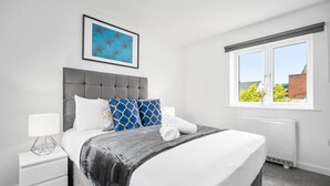 Un dormitorio moderno con un cabecero tapizado en gris, almohadas estampadas en blanco y azul, una cama impecablemente hecha con una manta gris, una mesita de noche blanca con una lámpara, y una ventana con vistas a edificios y árboles.