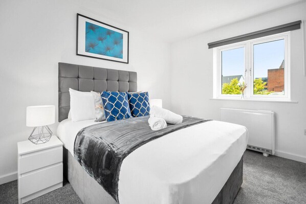 Ein modernes Schlafzimmer mit grauem gepolstertem Kopfteil, Kissen mit blau-weißem Muster, einem ordentlich bezogenen Bett mit grauer Decke, einem weißen Nachttisch mit Lampe und einem Fenster mit Blick auf Gebäude und Bäume.