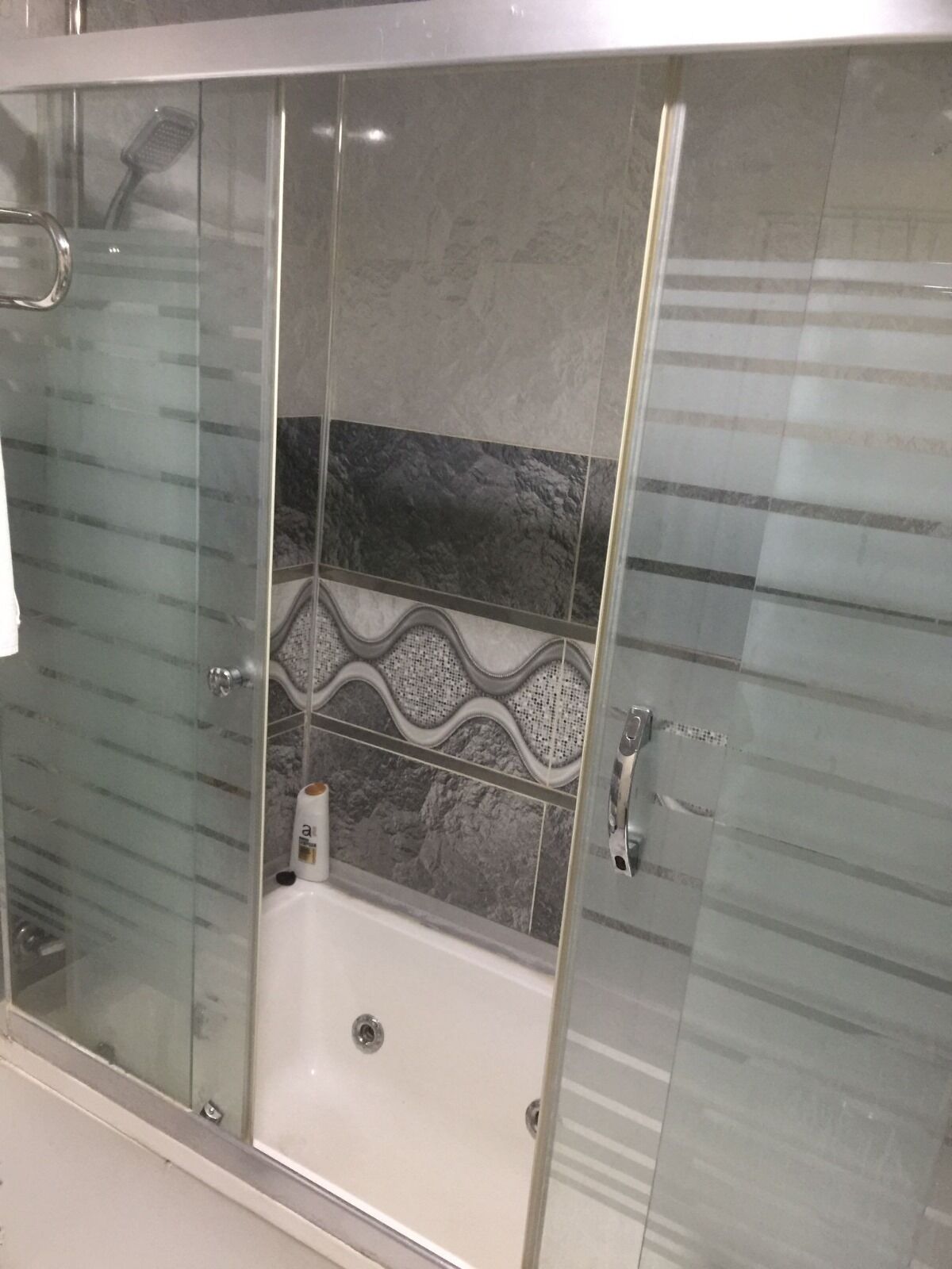 Une cabine de douche fermée par des parois en verre, avec une baignoire blanche, des carreaux aux motifs gris et blancs sur les murs, et une poignée métallique sur la porte.