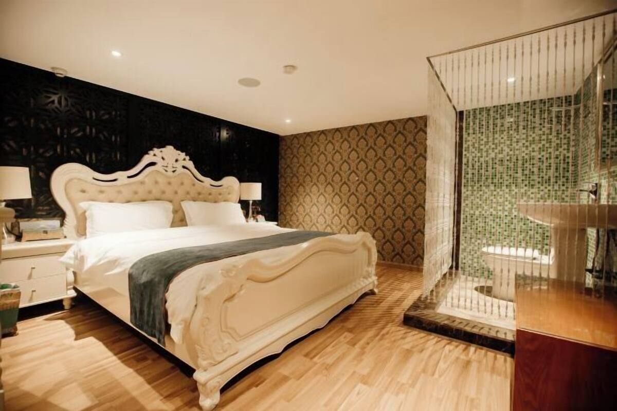 A large bed with a white headboard, a bedside table with a lamp, and a patterned wall.