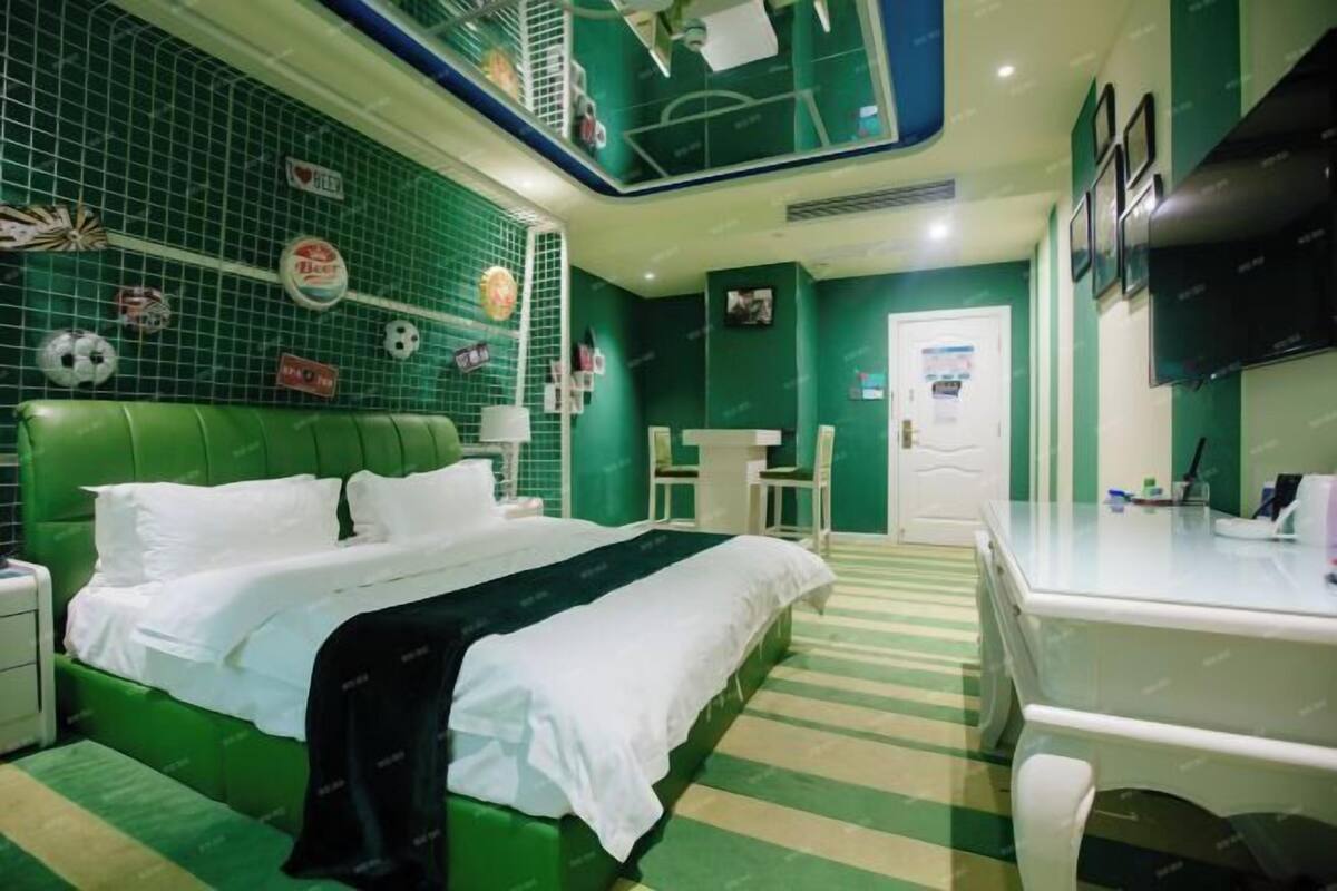 A hotel room with a green-striped floor, a bed with white linens, a desk with a chair, and a television on the wall.