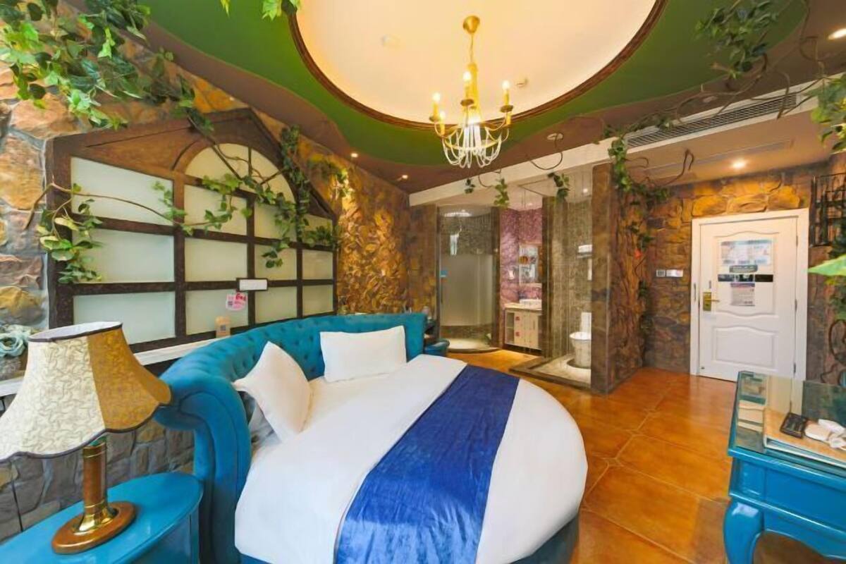 A bedroom with a large bed, a blue headboard, a chandelier, and a stone wall with plants.