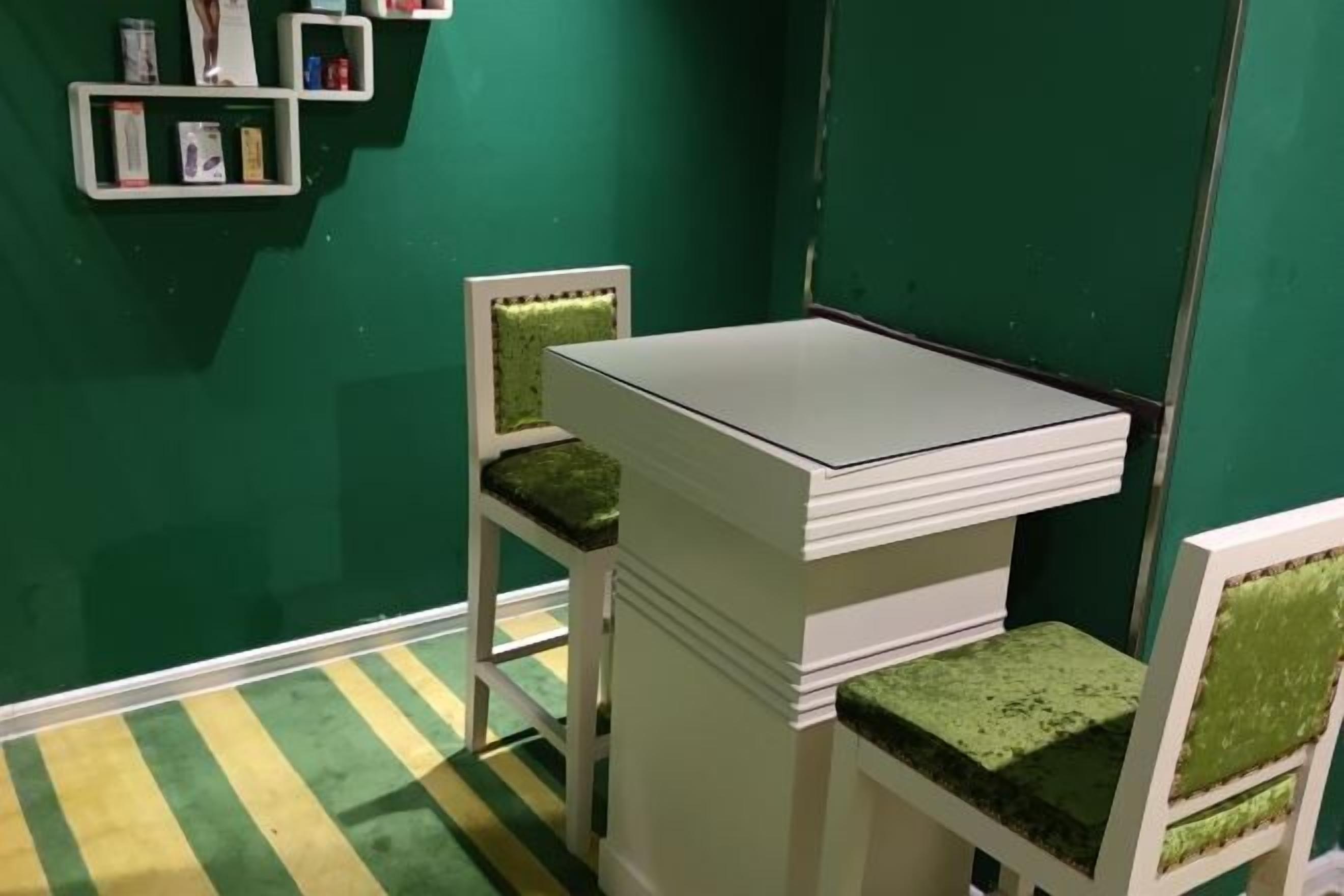 Una habitación pequeña con una mesa blanca y dos sillas con cojines verdes. Hay estantes con libros y un cuadro en la pared.