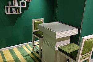 Una habitación pequeña con una mesa blanca y dos sillas con cojines verdes. Hay estantes con libros y un cuadro en la pared.