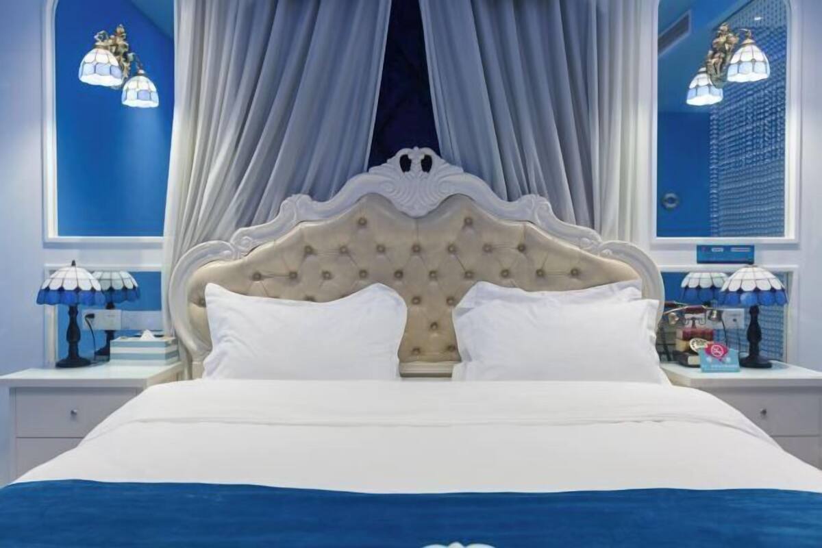 A bed with a tufted headboard, white bedding, and a blue accent at the foot of the bed. There are two bedside tables with lamps, and a window with curtains.