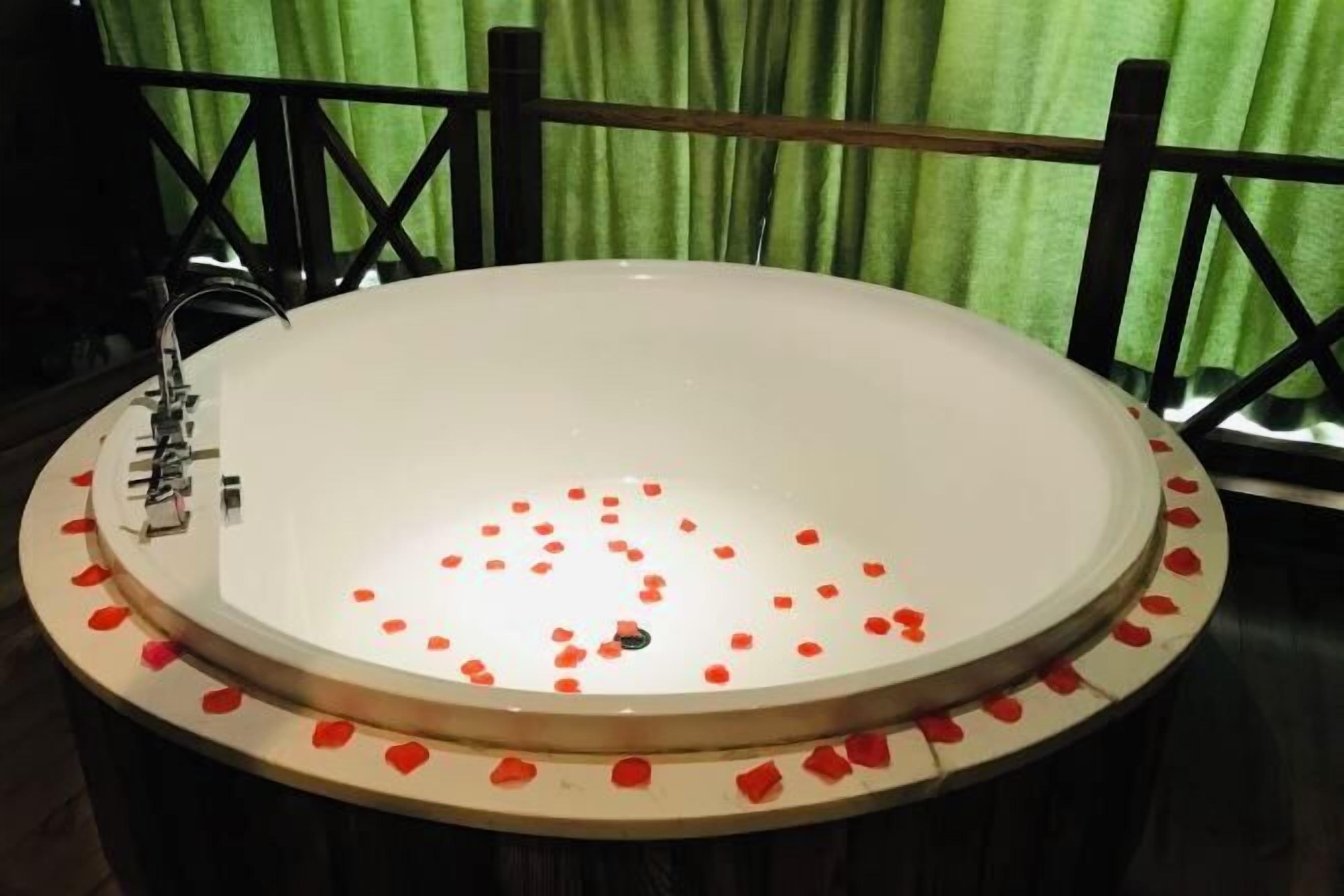 Una bañera redonda con pétalos de rosa, un grifo y una cortina verde al fondo.