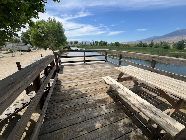 A wooden deck with picnic tables overlooking a body of water, surrounded by trees and a mountainous landscape.