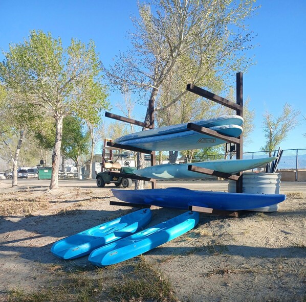 Kayaks and paddleboards stacked on a rack outdoors with trees and a clear sky in the background.