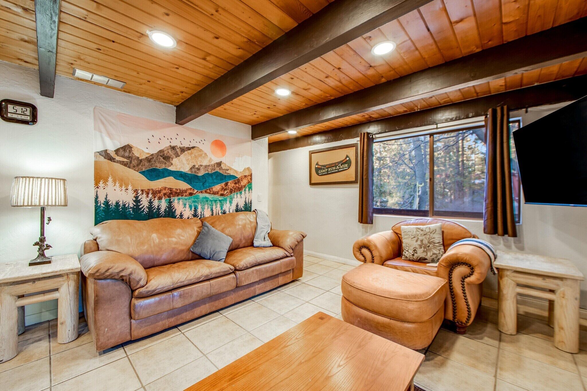 A cozy living room with a wooden ceiling, a mountain mural, a leather sofa, and a window with a view of trees.