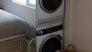 Een kleine wasruimte met een voorlader wasmachine en droger, een raam, een bed en een klein rieten mandje.