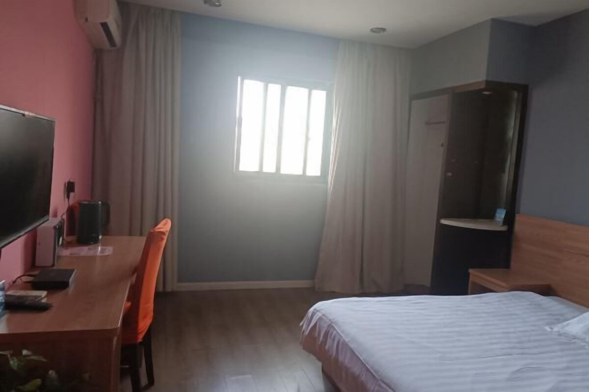 A hotel room with a bed, a desk with a chair, a television, a window with curtains, and a wardrobe.