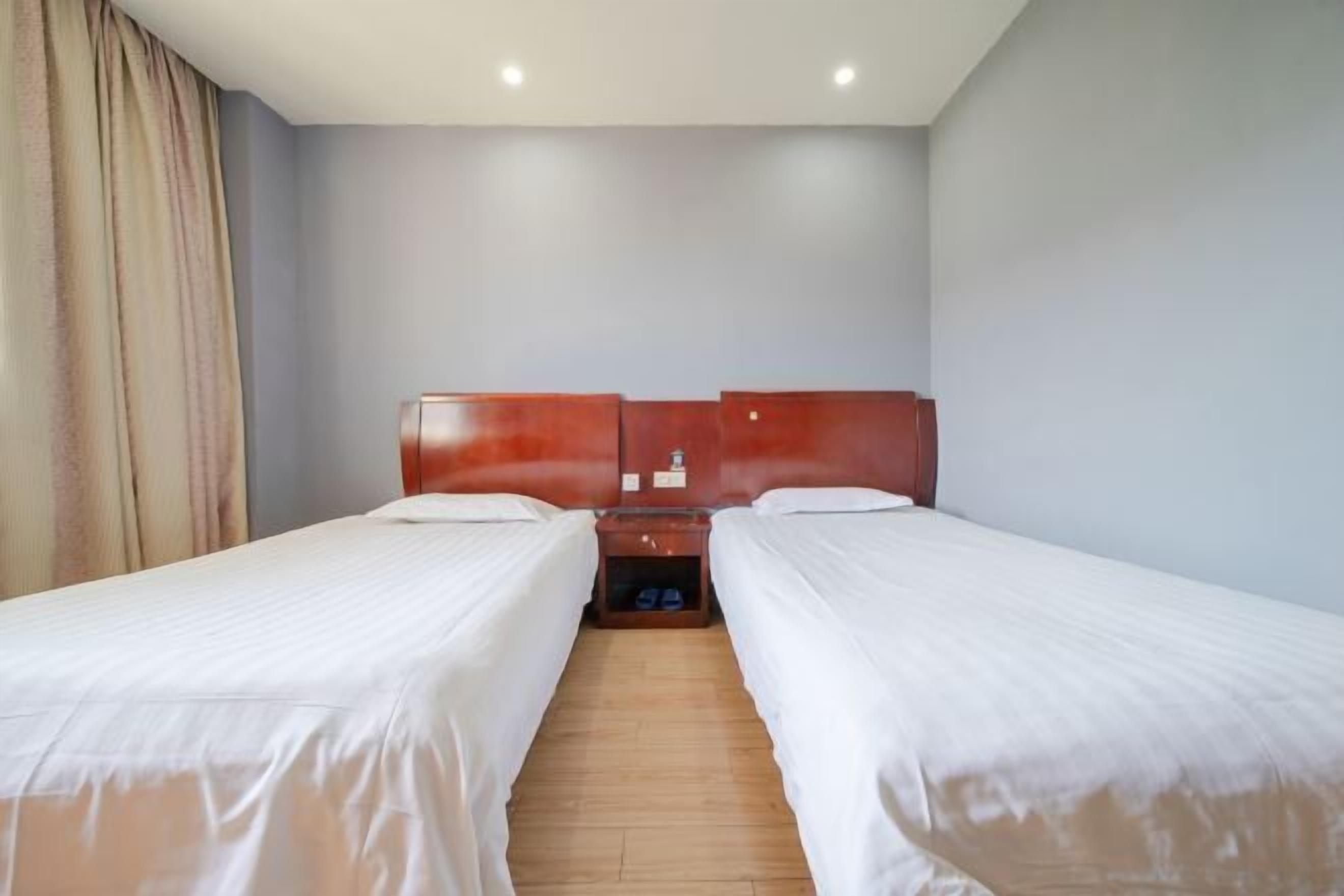 Habitación de hotel con dos camas individuales, cabeceros de madera, una mesita de noche con lámpara y suelo de madera.