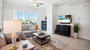 A modern living room with a grey sofa, a coffee table, a TV, and a ceiling fan. - Harbor Condos 305 - Brand new, steps to beach w/pool! (Madeira Beach)