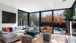 Un salon moderne avec un canapé gris, un pouf bleu et une table basse. Une grande fenêtre offre une vue sur une piscine et des arbres.