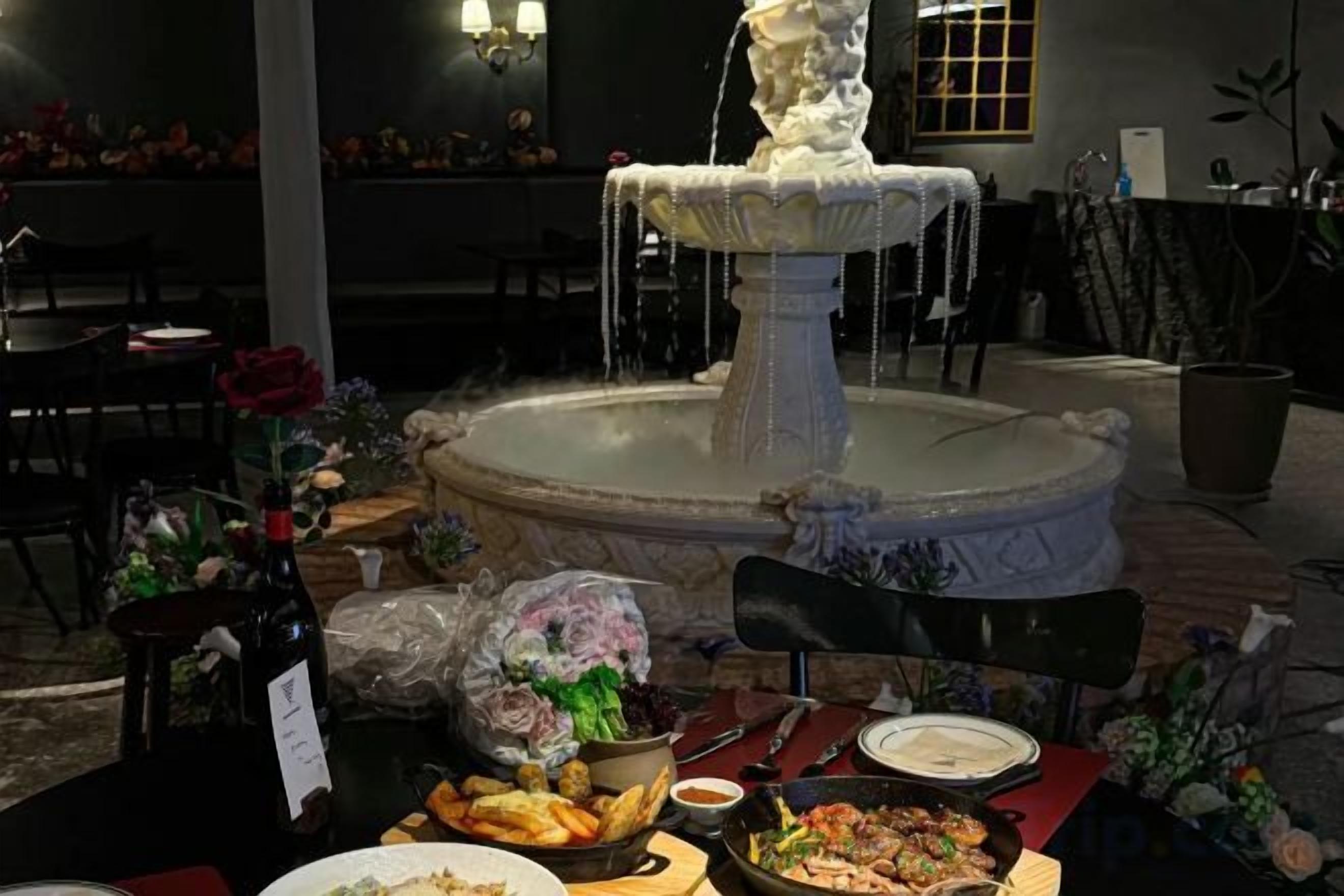 A dining area with a fountain centerpiece, tables set with plates and glasses, and a bottle of wine.