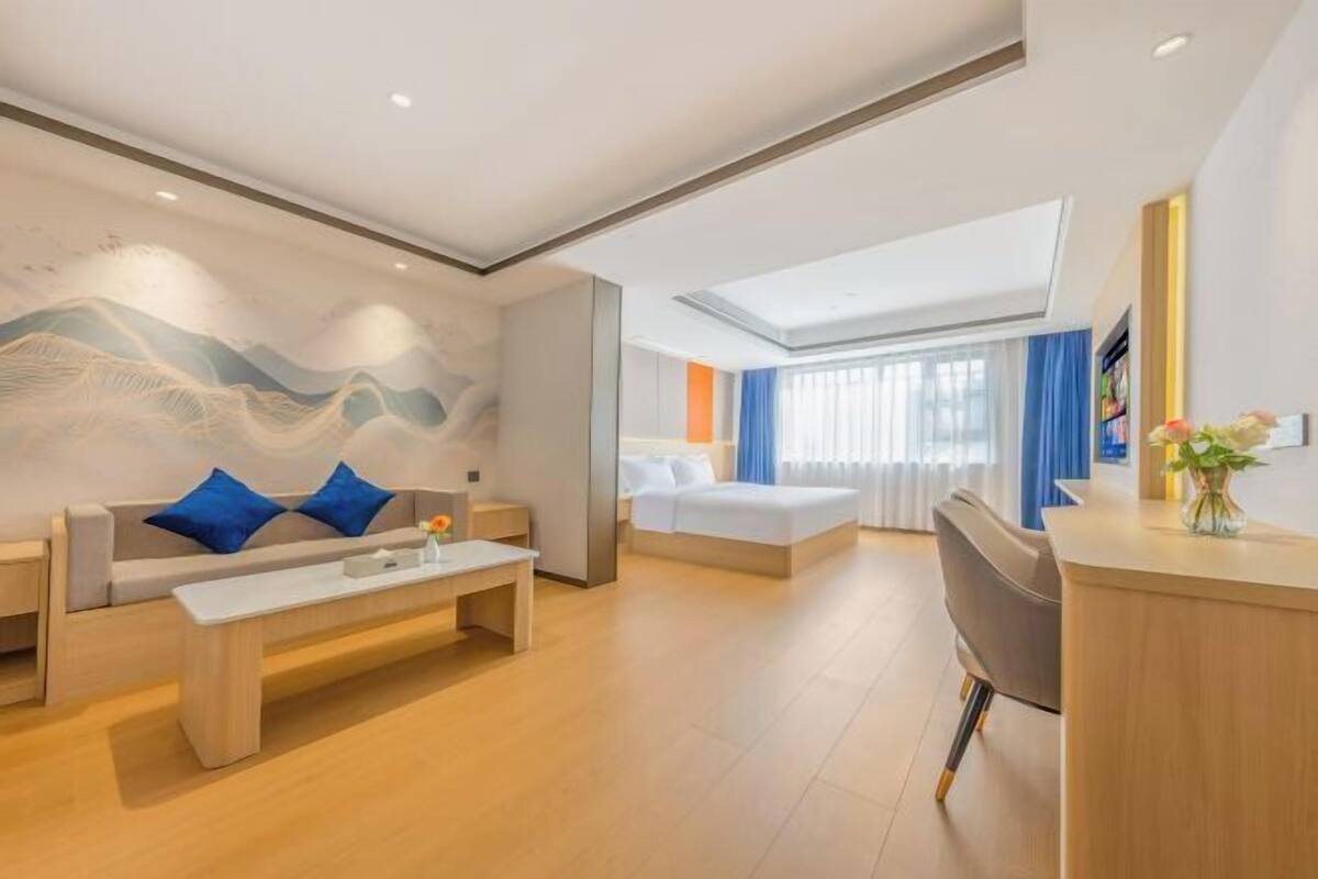 A modern hotel room with a sofa, a desk, a bed, and a large window.