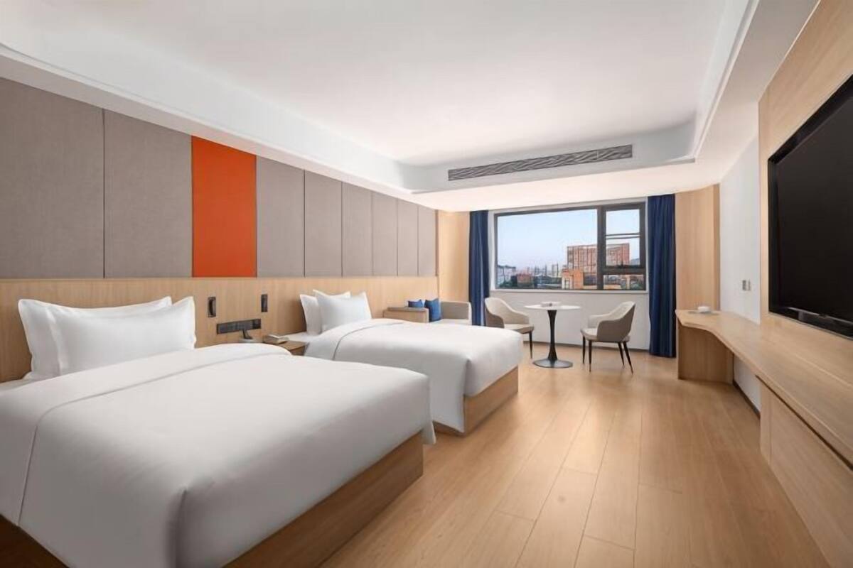 A hotel room with two beds, a TV, a desk, and a view of the city.