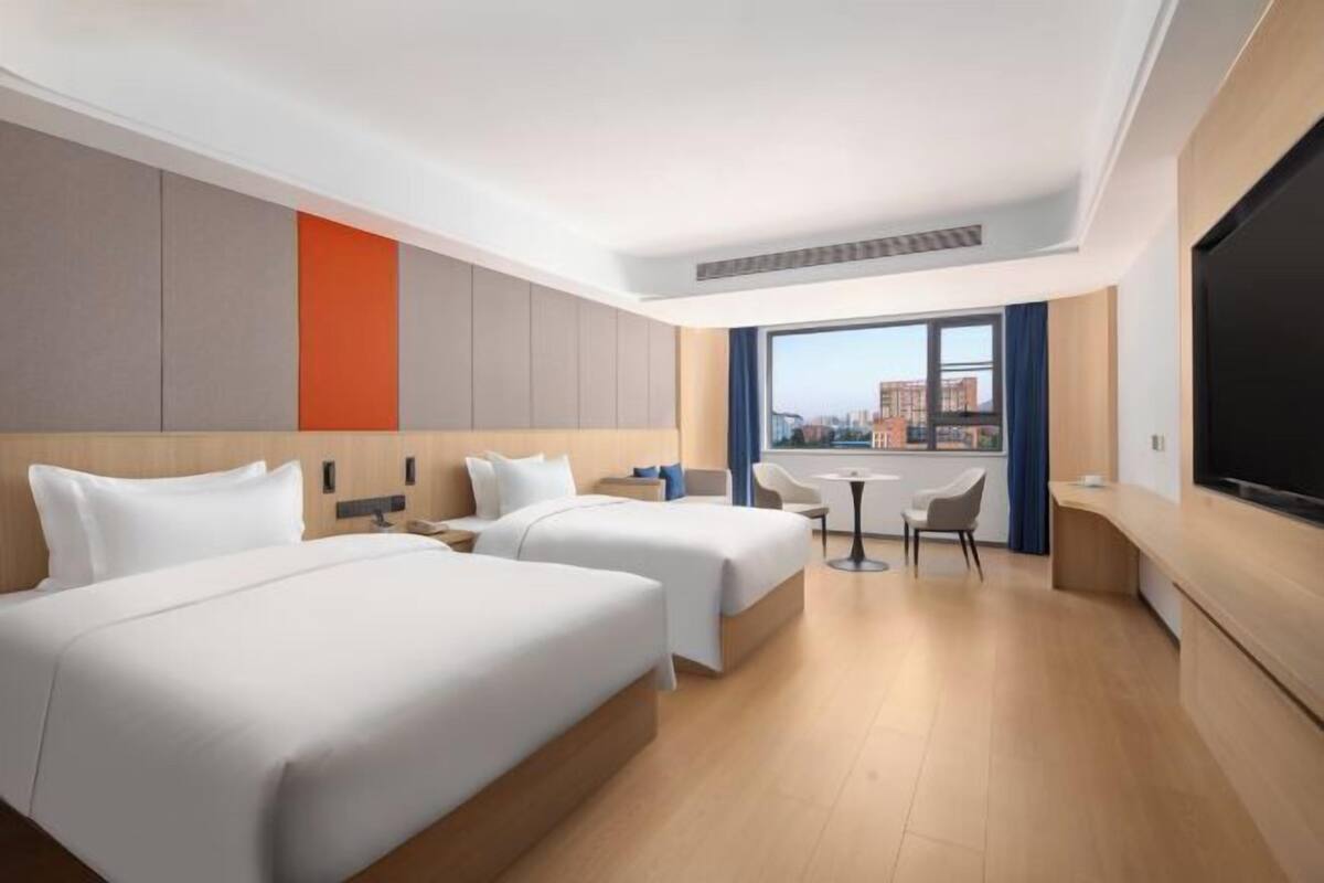A hotel room with two beds, a desk, a chair, a TV, and a large window with a city view.