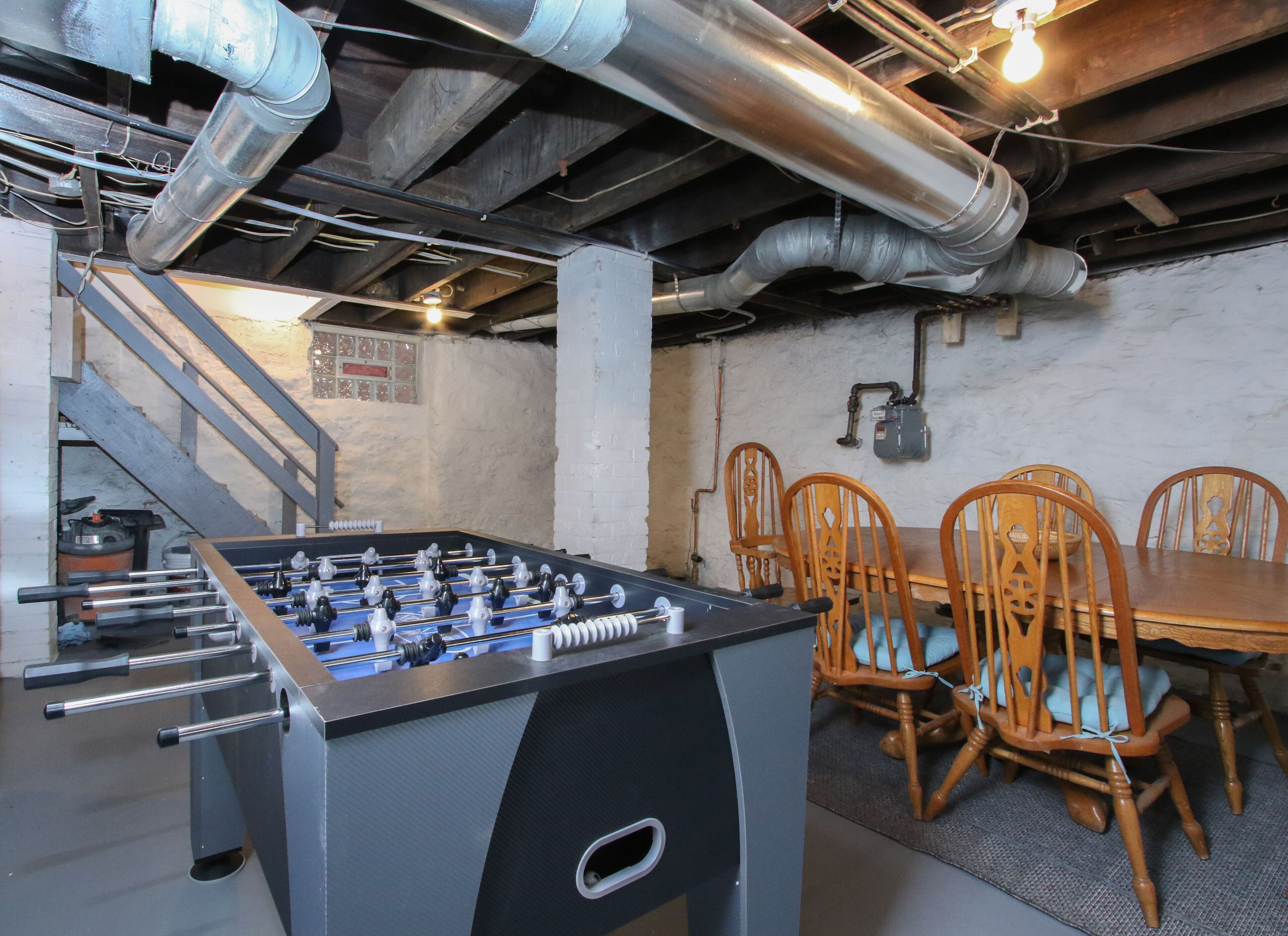 A foosball table in a room with a staircase, wooden chairs, and visible plumbing.