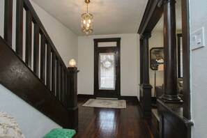 Un escalier en bois surmonté d’un lustre, une porte en bois sombre ornée d’un rideau en dentelle et un tapis menant à une pièce.