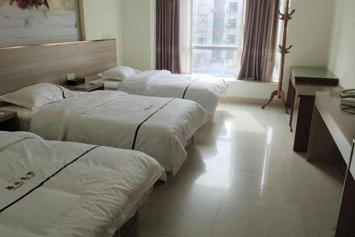 Habitación de hotel con tres camas, cabecera de madera, escritorio, silla y ventana con cortinas.