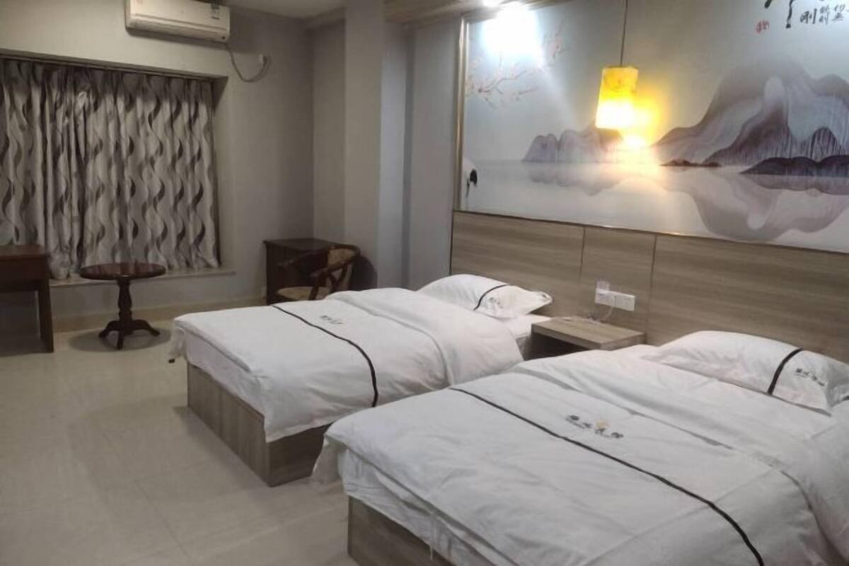 Habitación de hotel con dos camas, un escritorio, una silla, aire acondicionado instalado en la pared y un cuadro de montañas.