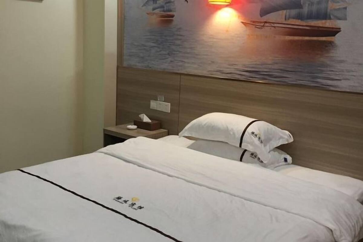 Habitación de hotel con cama, mesita de noche, un cuadro de barcos en el agua y cabecera de madera.