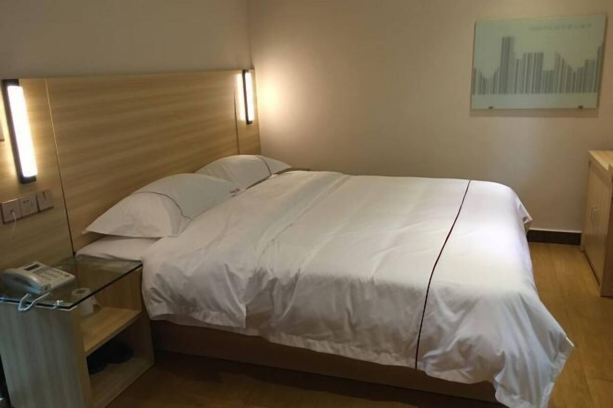 Una habitación de hotel con cabecera de madera, cama con sábanas blancas, mesita de noche con lámpara y un cuadro colgado en la pared.