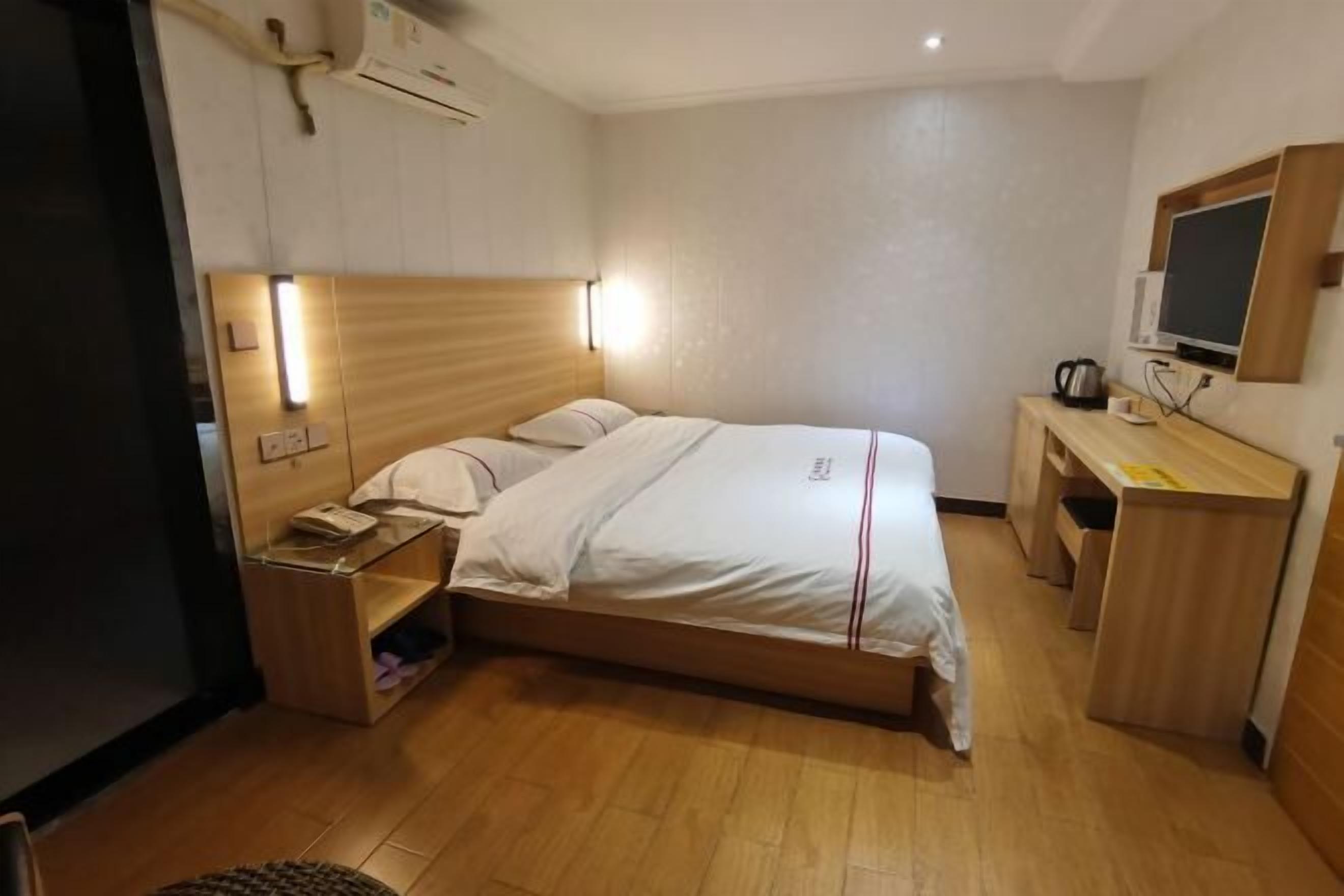 A hotel room with a wooden bed, bedside tables, a desk with a chair, a flat-screen TV, and an air conditioning unit.