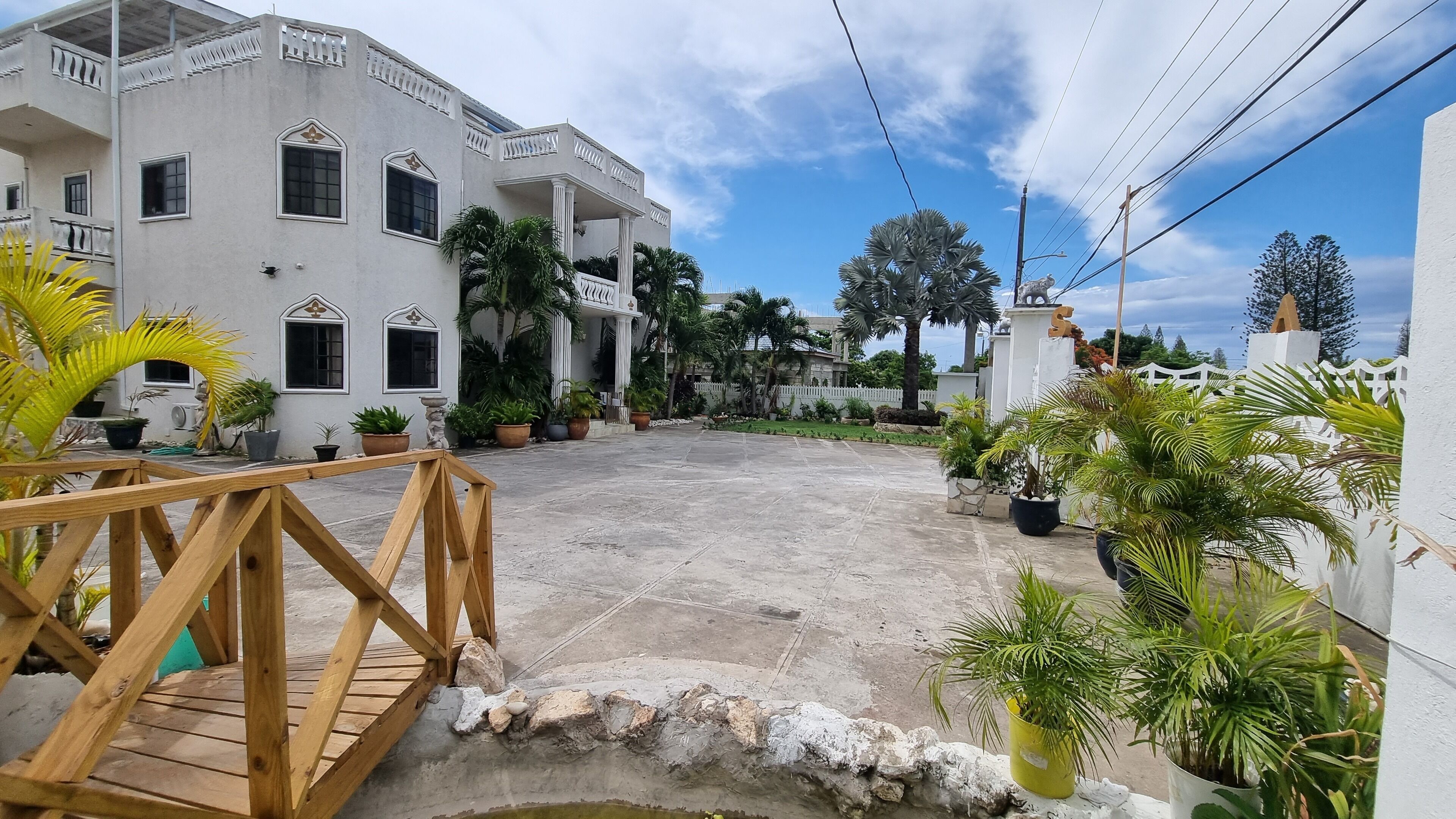 A spacious courtyard with a wooden bridge, surrounded by potted plants and a white building with balconies.