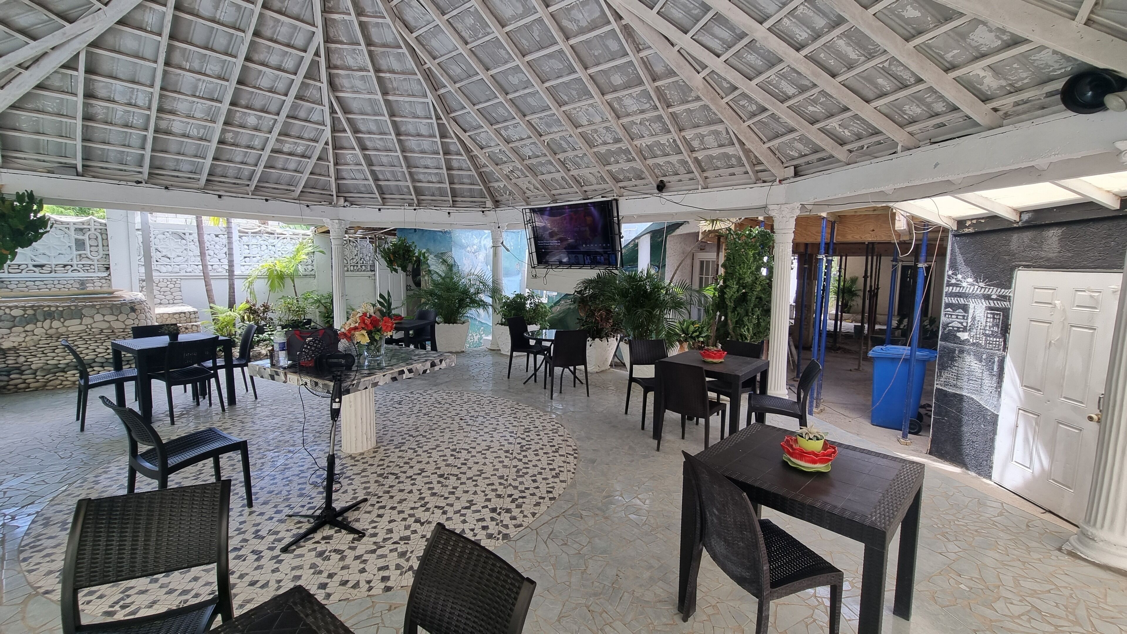 A covered outdoor seating area with tables and chairs, surrounded by potted plants and a tiled floor.