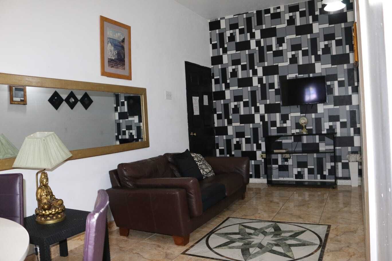 Una sala de estar con un sofá de cuero marrón, una mesita con una lámpara, una estatua decorativa de Buda, un cuadro enmarcado en la pared y una pared de azulejos con un diseño geométrico.