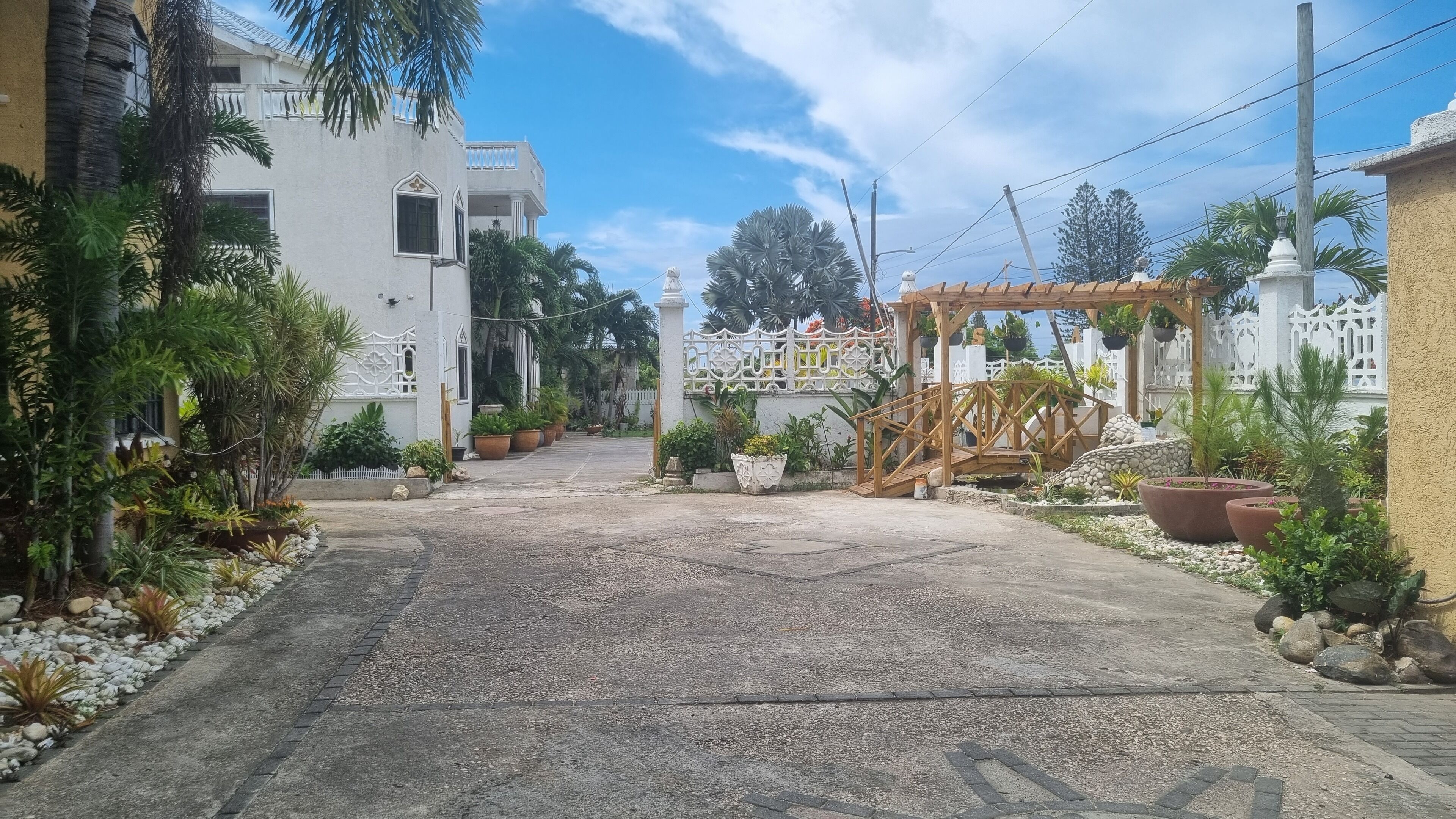 Una entrada con reja y un arco de madera, rodeada de plantas en macetas y un camino de grava que lleva a un edificio blanco con balcón.
