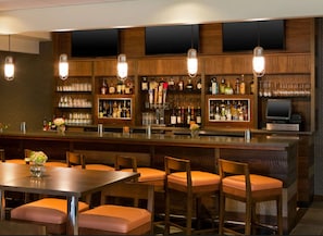Zona de bar con paneles de madera, varias botellas de licor, vasos y una pantalla de televisión. Hay taburetes de bar y una mesa con un jarrón.