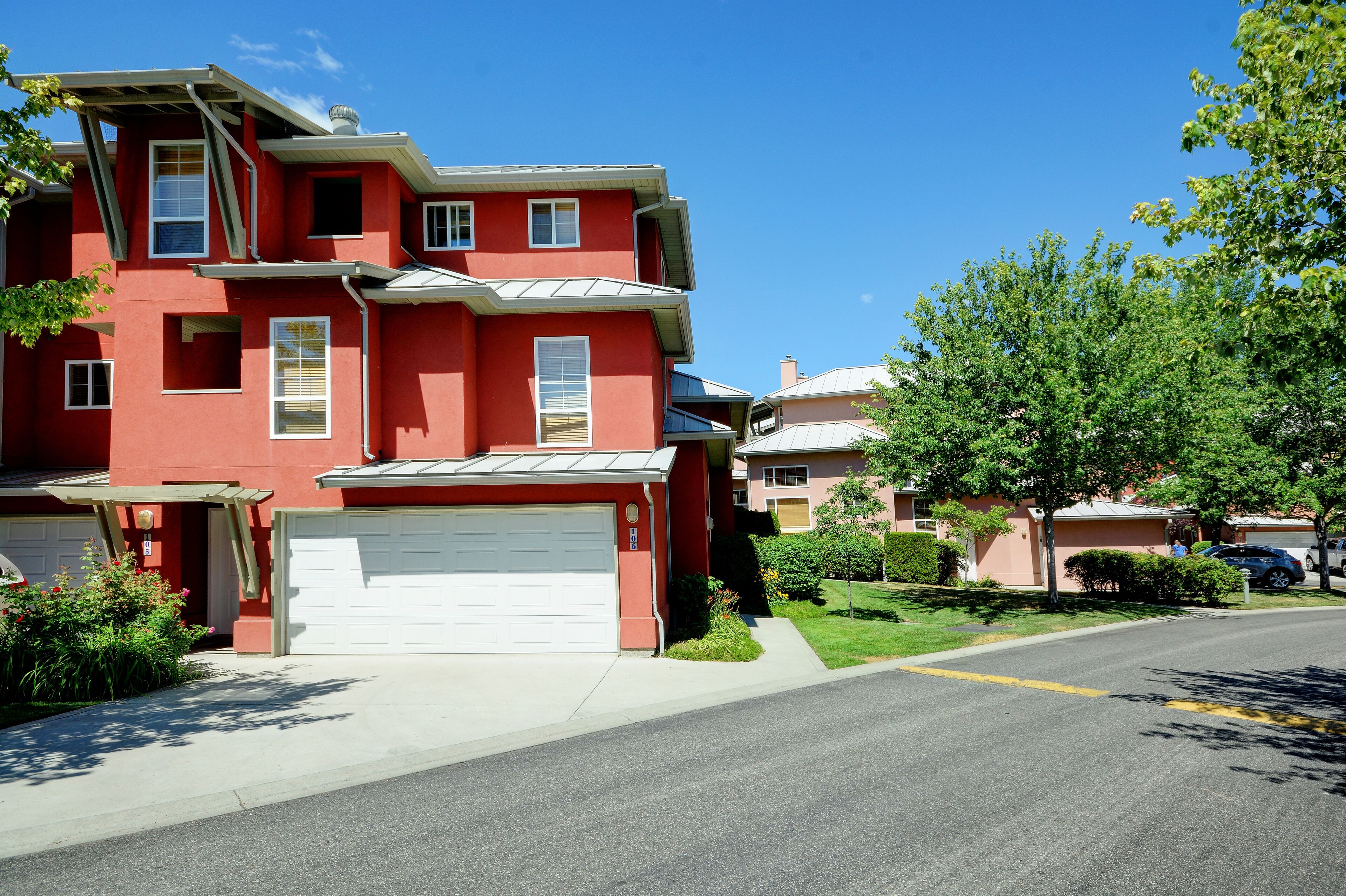 Une maison rouge à deux étages, avec une porte de garage blanche, entourée d’une pelouse bien entretenue et sous un ciel bleu dégagé.